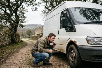Homme inspectant un van blanc d'occasion garé sur un chemin de gravier en milieu rural, examinant la carrosserie avec attention